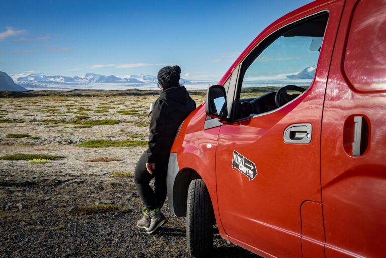 Person enjoying a scenic road trip in Iceland, standing beside a red van with mountains in the backdrop.